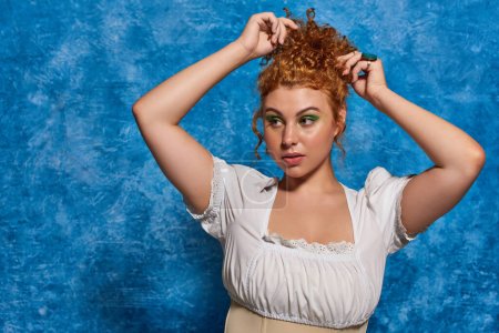 stylish curvy woman in white blouse adjusting red hair on blue textured backdrop, plus size fashion