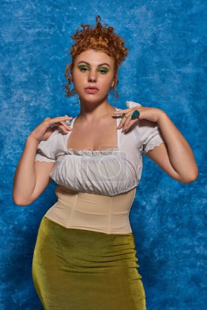 stylish and redhead plus size woman in white blouse looking at camera on blue textured backdrop
