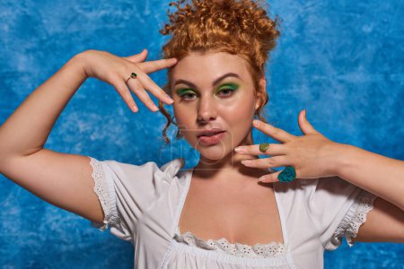 headshot of redhead plus size model in stylish attire posing with hands near face on blue backdrop