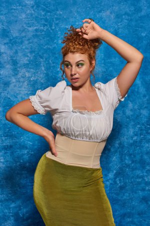 young redhead plus size woman in stylish clothes posing with hand on hip on blue textured backdrop