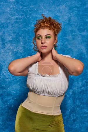 elegant redhead plus size woman in white blouse with hands behind neck looking away on blue backdrop