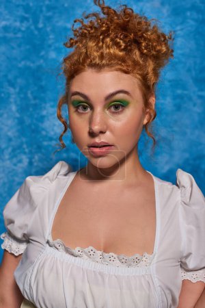 portrait of charming redhead woman in white blouse looking at camera on blue textured backdrop