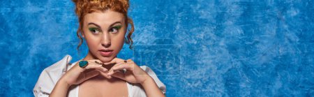 young redhead plus size model with hands near chin looking away on blue backdrop, horizontal banner