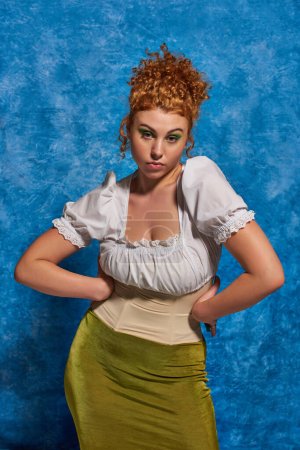 fashionable plus size model with red wavy hair posing with hands on hips on blue uneven backdrop