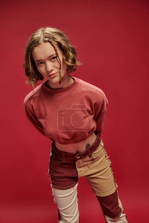 Persönlicher Stil, junge Frau in Patchworkhosen und abgeschnittenem Langarm posiert auf rotem Hintergrund