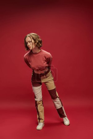 estilo personal, chica bonita joven en pantalones patchwork y manga larga recortada posando sobre fondo rojo