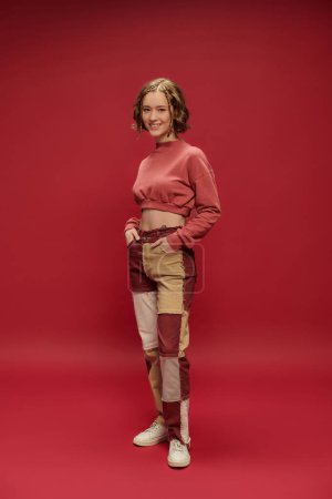 full length of happy girl in patchwork pants and cropped long sleeve with hands in pockets on red