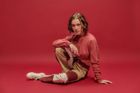Selbstdarstellung, junge Frau in Patchworkhosen und abgeschnittenem Langarm sitzt auf rotem Hintergrund