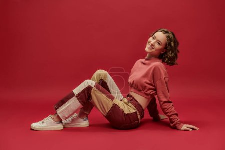expresión personal, mujer positiva en pantalones patchwork y manga larga recortada sentado sobre fondo rojo