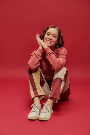 joyful woman in patchwork pants and cropped long sleeve sitting on red backdrop, youth culture