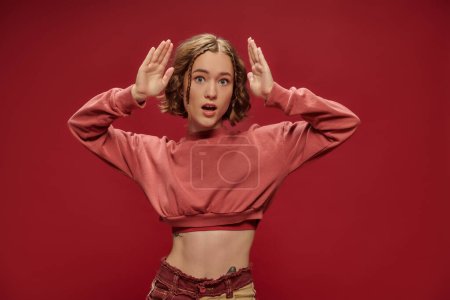 Photo for Shocked tattooed young woman with short hair and braids standing with open mouth on red backdrop - Royalty Free Image
