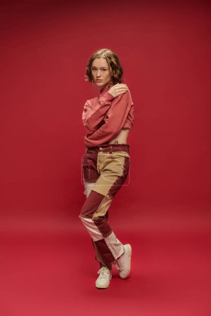 Foto de Estilo personal, mujer sensual en pantalones patchwork y manga larga recortada abrazando hombro en rojo - Imagen libre de derechos