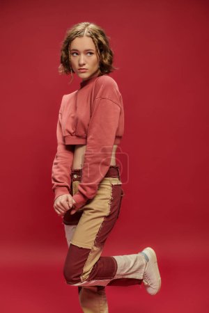 youth style, shy pretty woman in patchwork pants and cropped long sleeve posing on red backdrop