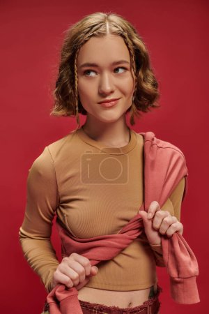 junge hübsche Frau in Patchwork-Hosen bindet Pullover über abgeschnittenem Langarm auf rotem Hintergrund, Stil