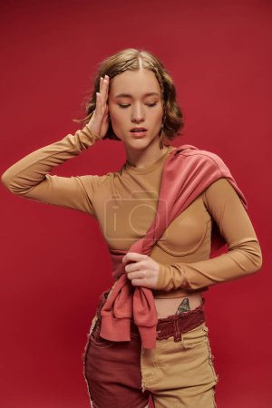 young stylish woman in patchwork pants posing in tied jumper over long sleeve on red backdrop