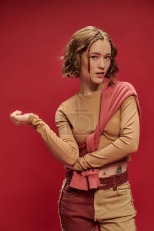 trendige junge Frau in Patchworkhosen posiert im Pullover über abgeschnittenem Langarm vor rotem Hintergrund