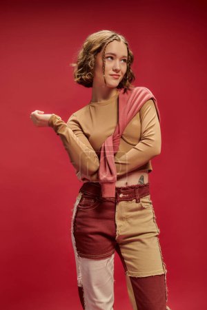 fröhliche junge Frau in Patchworkhosen posiert im Pullover über abgeschnittenem Langarm vor rotem Hintergrund