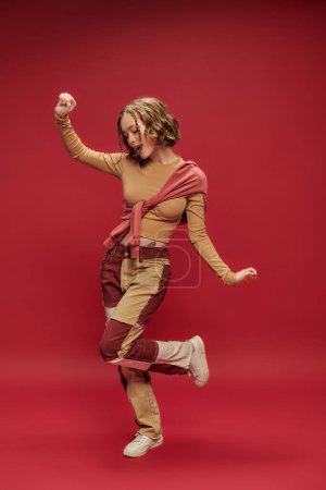 fröhliche junge Frau in Patchworkhosen posiert im Pullover über abgeschnittenem Langarm vor rotem Hintergrund