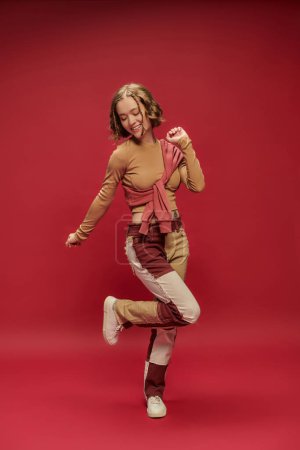 Foto de Mujer joven excitada en pantalones patchwork posando en jersey sobre manga larga recortada sobre fondo rojo - Imagen libre de derechos