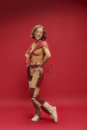 positive young woman in patchwork pants posing in jumper over cropped long sleeve on red backdrop