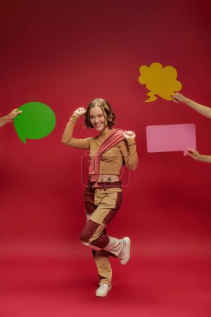 Foto de Mujer alegre en pantalones patchwork y puente sobre manga larga bailando cerca de personas con burbujas del habla - Imagen libre de derechos