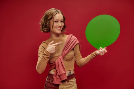 glückliche Frau in Patchworkhose und Pullover über abgeschnittenem Langarm, die auf Sprechblase auf Rot zeigt
