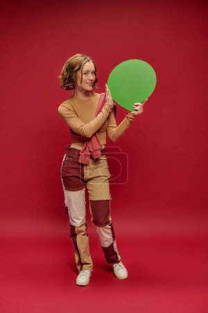 junge Frau in Patchworkhose und Pullover über abgeschnittenem Langarm zeigt auf Sprechblase auf Rot