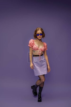 Foto de Bonita mujer joven en top recortado con corazones y falda posando en gafas de sol en púrpura - Imagen libre de derechos