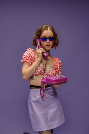pretty girl in trendy sunglasses and cropped top with hearts talking on retro telephone on purple