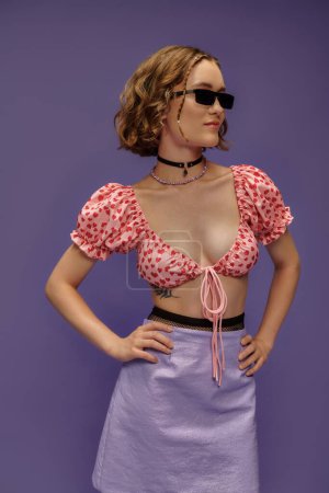 Téléchargez les photos : Jeune femme confiante dans des lunettes de soleil à la mode et haut recadré posant avec les mains sur les hanches sur violet - en image libre de droit