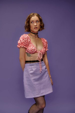 mujer joven pensativa en top recortado y falda posando sobre fondo púrpura, estilo personal