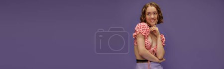 smiling young woman in cropped top posing with hand near chin on purple background, banner