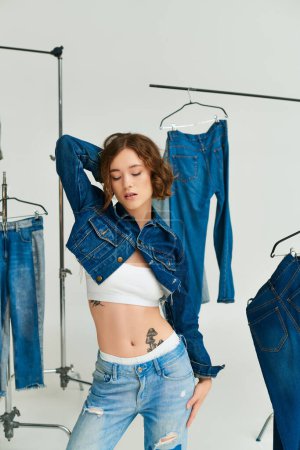 Photo for Charming young woman with tattoo posing in cropped blue jacket among denim clothes on grey backdrop - Royalty Free Image