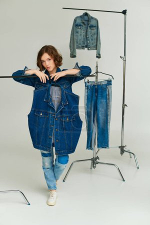 Jeansmode, hübsche junge Frau posiert neben trendiger blauer Weste zwischen Jeans auf grauem Hintergrund