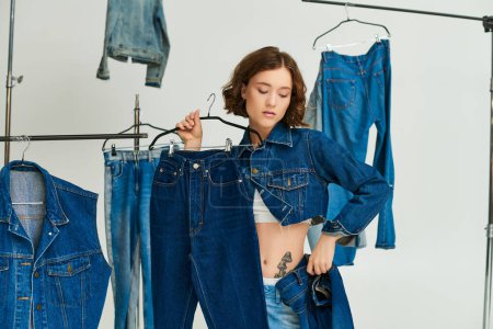 Photo for Tattooed young woman in blue cropped jacket holding jeans among denim clothes on grey backdrop - Royalty Free Image