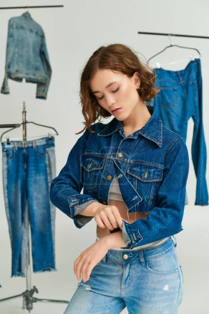 young model adjusting sleeve on cropped jacket and posing among denim clothes on grey backdrop