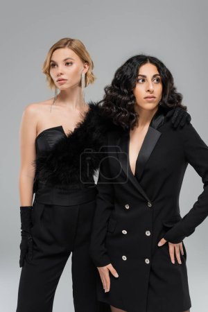 brünette und blonde multirassische Freundinnen in total schwarzer, eleganter Kleidung posieren vor grauem Hintergrund