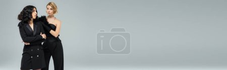 Téléchargez les photos : Femme blonde avec brune multiraciale petite amie en noir élégant porter sur gris, bannière de mode - en image libre de droit