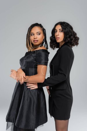 Vielfalt und Mode, zwei multiethnische Freundinnen in schwarzer eleganter Kleidung posieren vor grauem Hintergrund