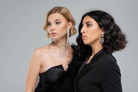 Photo for Portrait of multiracial girlfriends in black elegant attire and silver earrings on grey backdrop - Royalty Free Image