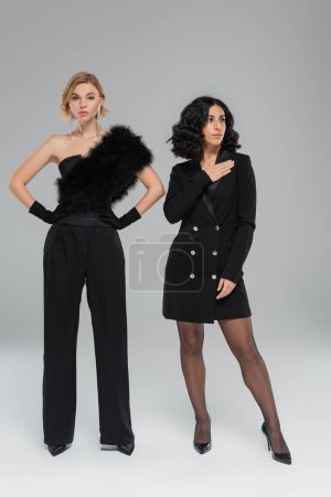 Photo for Blonde woman with hands on hips near trendy multiracial girlfriend in black on grey, full length - Royalty Free Image