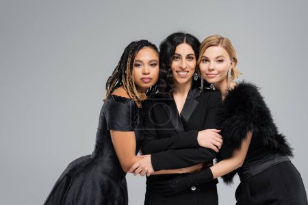 Photo for Happy multiracial girlfriends in black elegant outfits embracing on grey, diversity and fashion - Royalty Free Image