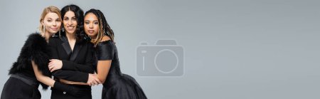 Photo for Joyful multiracial girlfriends in black elegant attire embracing on grey, horizontal banner - Royalty Free Image