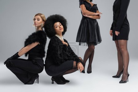 Blonde und brünette multiethnische Freundinnen in schwarzer modischer Kleidung sitzen auf grauem Hintergrund