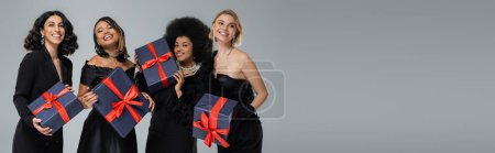 Photo for Joyful multiethnic fashionistas in total black elegant clothing posing with presents on grey, banner - Royalty Free Image