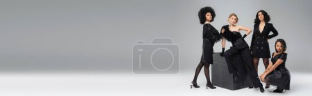 Photo for Multicultural girlfriends in dark elegant attire posing near black cube on grey, horizontal banner - Royalty Free Image