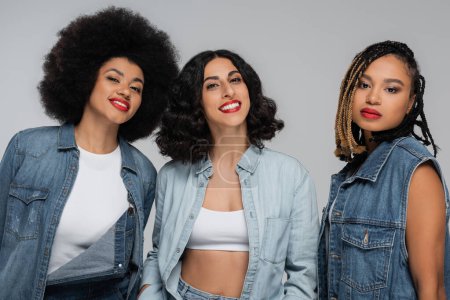 fröhliche multirassische Freundinnen in trendiger Jeans-Kleidung lächeln vor grauer Kulisse im Studio in die Kamera