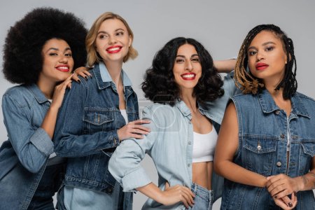 attraktive und glückliche multiethnische Freundinnen in Jeans posieren für Grau, Vielfalt und Mode
