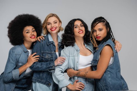 glückliche multiethnische Freundinnen in trendiger Jeanskleidung, die in die Kamera schauen, während sie auf grau posieren
