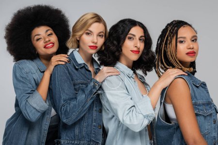 multirassische Freundinnen in Jeans tragen Händchen auf Schultern und lächeln in die Kamera